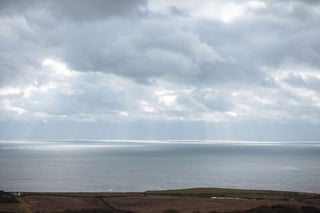 Shafts of light break through heavy cloud over a calm sea off the Isle of Wight coast, farmland in the foreground.