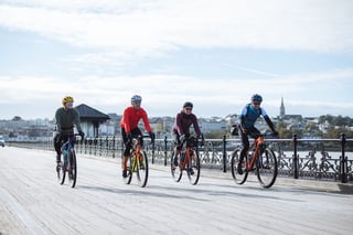 Four gravel riders cycle along an Isle of Wight seafront pier past ornate iron railings, the town behind.