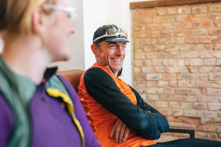 A rider in an orange gilet and cap laughs while chatting in a café, another rider blurred in the foreground.