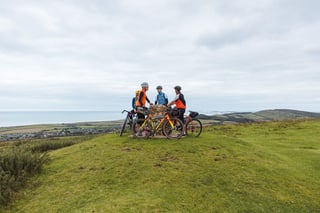 Riders pause with their bikes around a stone trig point on a grassy hilltop, the sea and coast beyond.