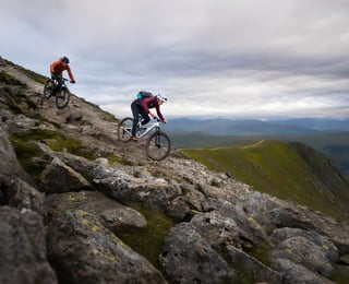 Two Rider Resilience mountain bikers descend a rocky ridge track above a green valley under heavy cloud.