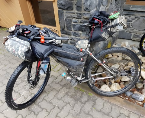 Tour Divide bikepacking bike loaded with frame, bar and seat bags, parked on paving against a stone wall.