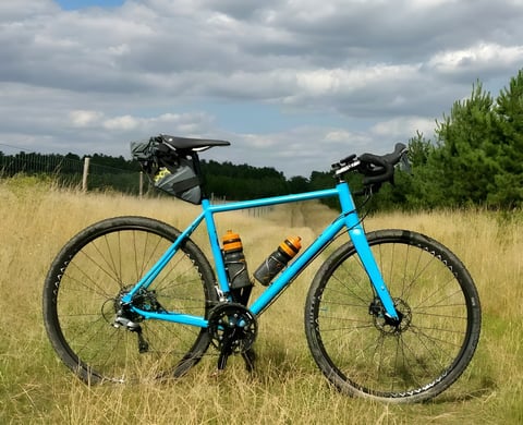 A blue gravel bike with drop bars and a seat pack stands in a grass field under a cloudy sky.