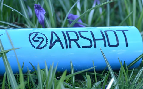 Close-up of the blue Airshot tyre inflator cylinder with its logo, lying in grass beside purple flowers.