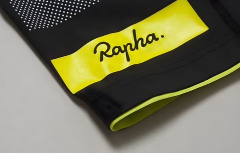 Close-up of a Rapha Brevet bib shorts leg hem with a yellow logo panel, white dots and yellow gripper edge on black fabric.