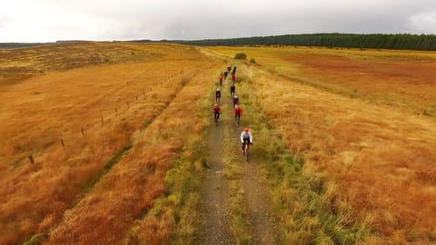Gravel Rocks Training Camp riders in a line along a grassy track across golden moorland, seen from above.