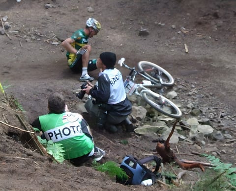 Mountain bike racer sits on the ground after a crash while two photographers crouch nearby and the bike lies on rocks.
