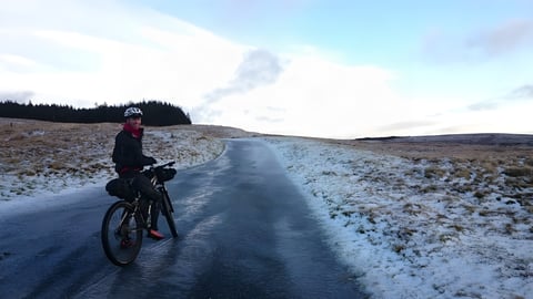 Bikepacker pauses on an icy road climbing towards Llangurig, snow on the moorland, for the Cafe Racer winter event.