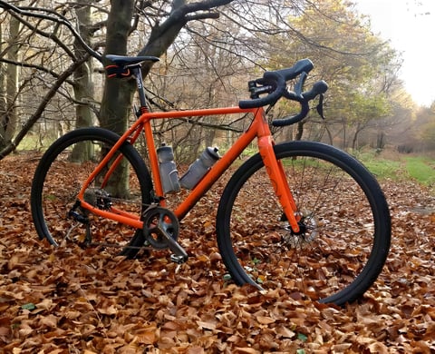 Orange Mason Bokeh gravel bike with two bottles, parked on autumn leaves in woodland.
