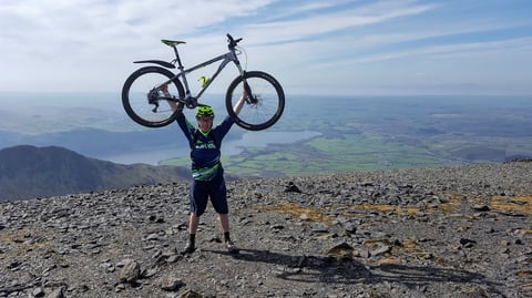 James Pearson holds his Whyte hardtail overhead on a stony Lake District summit, with a lake and fields far below.