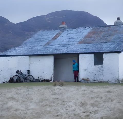 Bike leaning against a whitewashed Scottish bothy with a figure in the doorway beneath a hill.