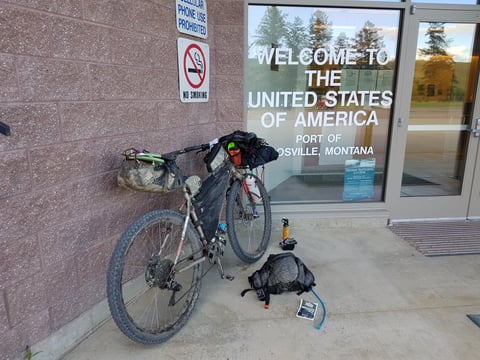 Mud-caked bikepacking bike outside the US port of entry at Roosville, Montana, with kit laid out on the ground.