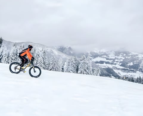 Fatbike rider in orange climbs a snowy slope above a valley on Stage 1 of the Snow Bike Festival in Switzerland.