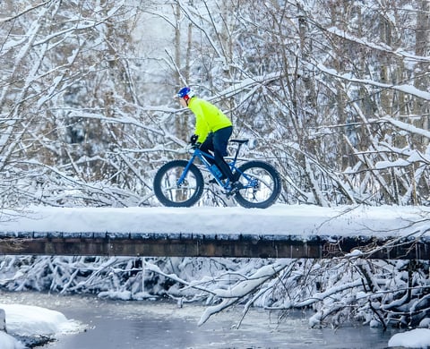 Rider in a yellow jacket crosses a snow-covered bridge on a blue fatbike during Stage 2 of the Snow Bike Festival.