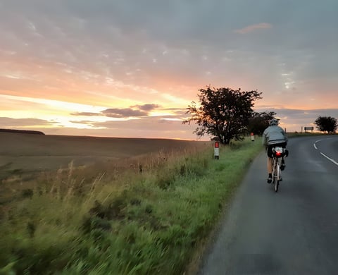 Cyclist with a rear light and race number rides a quiet country road at sunrise, an example of staying local on a bike.