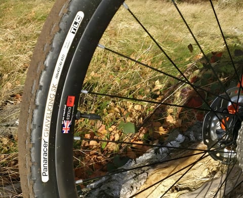 Panaracer GravelKing SK 43c TLC tyre on a black rim, caked in dry mud on a grassy trail.