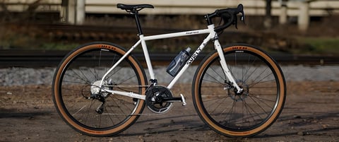White Surly Midnight Special steel road bike with tan-wall tyres and disc brakes, side view beside a railway.