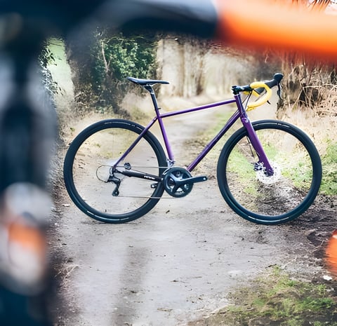 Purple Cotic Escapade 2018 steel gravel bike with yellow bar tape standing on a muddy country track.