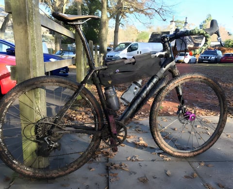Mud-spattered Cannondale Slate with a frame bag leaning against a wooden fence beside a car park.