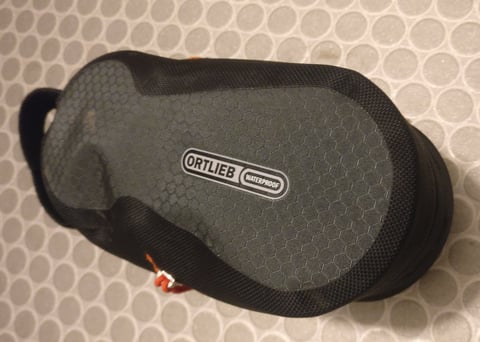 Top of the Ortlieb Cockpit Pack with its Waterproof badge on grey hexagon-pattern fabric, lying on a tiled floor.