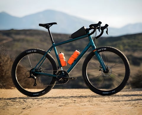 Salsa Journeyman gravel bike in teal with orange bottles, WTB Nano 27.5 tyres and a top tube bag, on dusty ground.