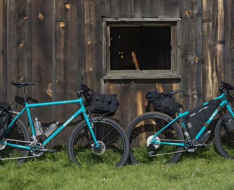 Two teal Surly Bridge Club bikes loaded with bikepacking bags, leaning against a weathered wooden barn.