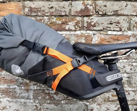 Ortlieb Seat-Pack in grey with orange straps mounted under a saddle against a brick wall.