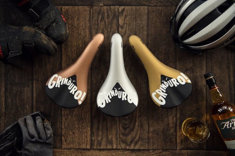 Three Fabric Grinduro saddles in copper, silver and gold, beside a bottle of Arran whisky and a glass.