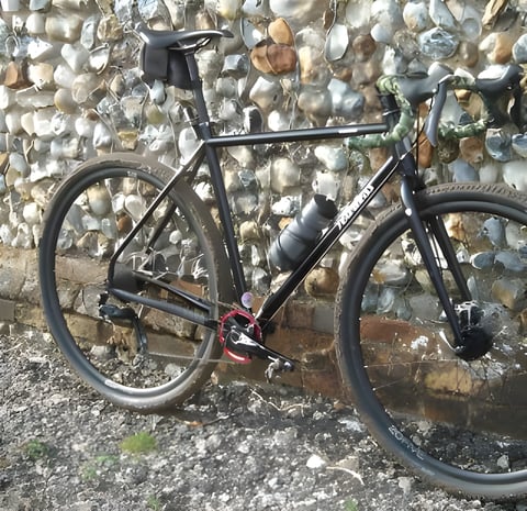 Black Fearless Vulture steel gravel bike leaning on a flint wall, with camo bar tape and a saddle bag.