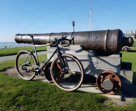 Bombtrack Audax steel road-plus bike with tan-wall tyres leaning on a cannon on a clifftop green.