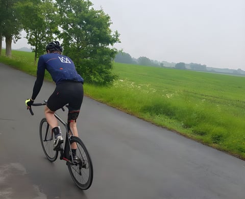 Cyclist in the navy Mason X Morvelo summer jersey rides away along a wet country lane beside a green field.