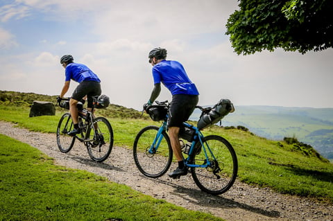 Two bikepackers in blue jerseys ride a gravel track above green hills on The Distance in the Peak District.