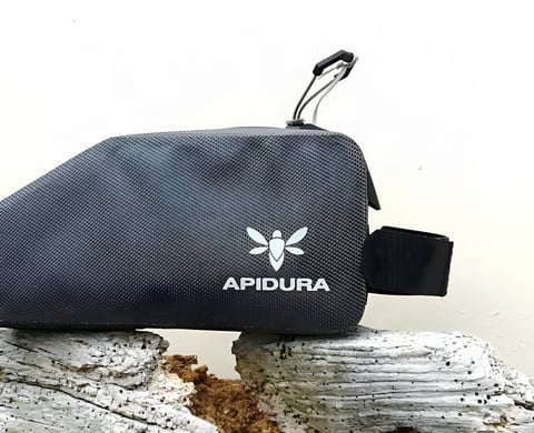 Apidura Expedition top tube bag in dark grey with silver logo, side view, resting on weathered wood.