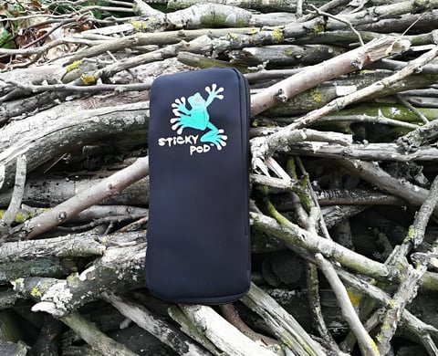 Black neoprene Large Sticky Pod with its green frog logo lying on a pile of lichen-covered sticks.