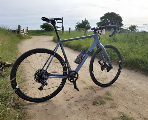 Lauf True Grit gravel bike in blue-grey stands on a dry Yorkshire farm track with a gate and grass verge behind.