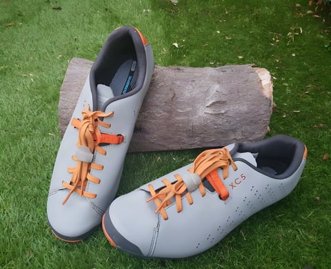Shimano XC5 SPD shoes in grey with orange laces, resting against a log on grass.