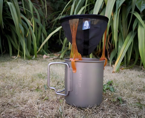 The GSI Outdoors Java Drip coffee filter set up over a titanium mug on grass.