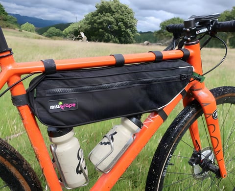 Miss Grape Internode frame bag strapped to the top tube of an orange gravel bike, with two bottles beneath.