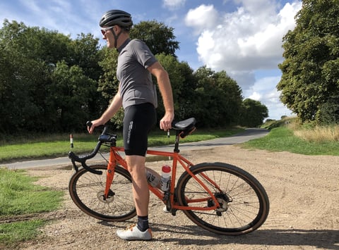 Rider in Endura FS260-Pro bibshorts stands beside an orange gravel bike on a gravel track by a country road.