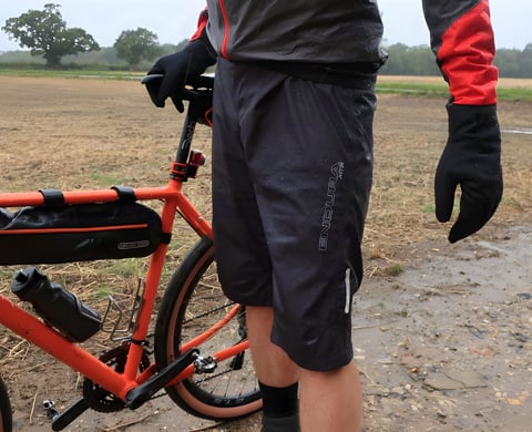 Rider in Endura MTR Waterproof Shorts and a rain jacket stands beside an orange gravel bike on a wet farm track.