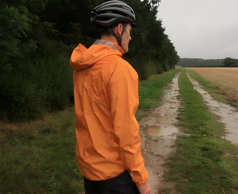 Rider seen from behind in the orange Endura MTR Shell Jacket on a puddled farm track in the rain.