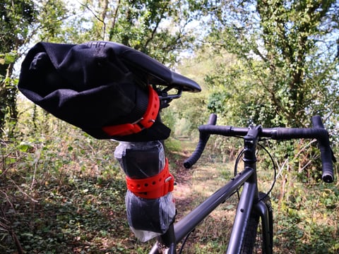 Voile straps securing a tool roll beneath the saddle and a spare tube on the seatpost of a gravel bike on a leafy lane.