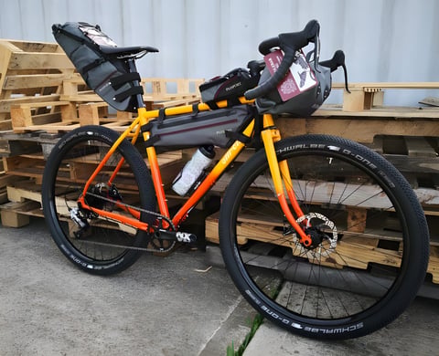 All-City Gorilla Monsoon in orange-to-yellow fade with Schwalbe G-One tyres and Passport bikepacking bags, against pallets.
