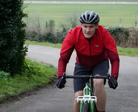 Rider in a red GORE R5 WINDSTOPPER long sleeve shirt and helmet rides a green gravel bike down a country lane.
