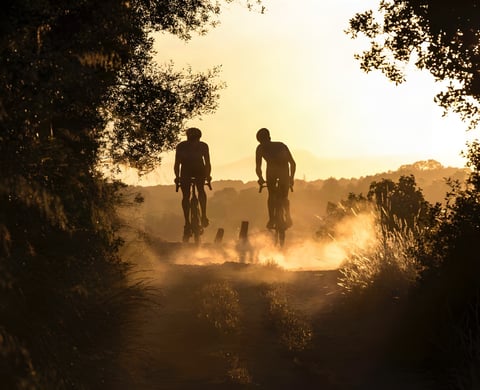 Two gravel riders silhouetted in dusty golden sunset light on a dirt track, promoting Toros de Gravel in Mallorca.