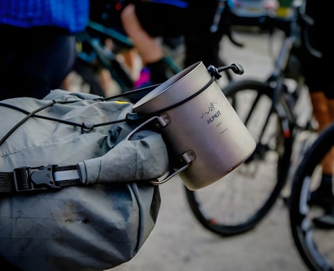Alpkit titanium mug strapped to a bikepacking bag, with gravel riders and their bikes gathered behind.