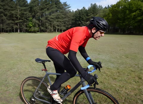 Rider in a red Le Col Therma Jersey and arm warmers pedals a gravel bike across a grassy clearing.
