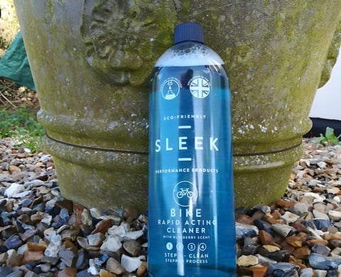 Bottle of Sleek Bike Rapid Acting Cleaner with blueberry scent standing against a mossy stone planter on gravel.