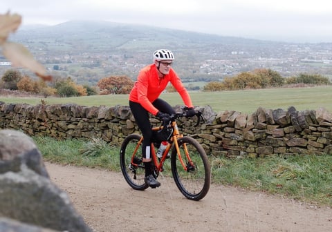 Rider in a bright orange POC AVIP Ceramic Thermal Jersey on a gravel bike beside a dry-stone wall.