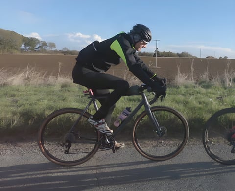 Rider in the black and hi-vis yellow Galibier Mistral Foul Weather Jacket on a gravel bike beside a Suffolk field.